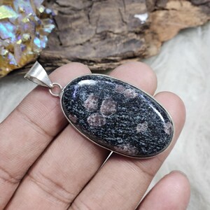 Peut inclure: Pendentif ovale avec une pierre sombre mouchetée sertie dans un cadre en métal argenté. La pierre a une base noire avec des taches brun rougeâtre. Le pendentif est suspendu à une bélière argentée.