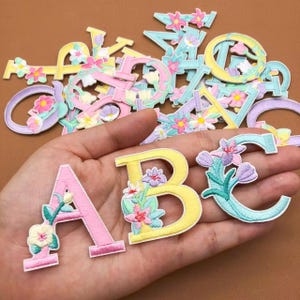 Könnte beinhalten: Gestickte Buchstaben mit Blumen in Pastellfarben. Die Buchstaben A, B und C sind im Fokus, gehalten in einer Hand. Weitere Buchstaben sind im Hintergrund verstreut. Jeder Buchstabe ist mit bunten Blumen verziert.