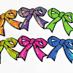 May include: Six colourful sequined bow patches in various colours including green, blue, pink, yellow, and purple. The bows are outlined in black.