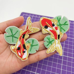 May include: Two embroidered iron-on patches featuring koi fish swimming in a pond with lily pads. The fish are red, white, and black with gold accents. The lily pads are green.