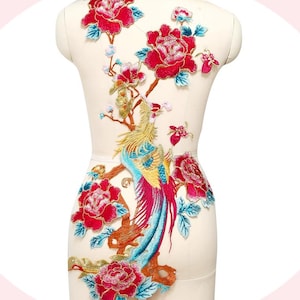 Könnte beinhalten: Gestickte Stoffapplikation mit einem Pfau- und Blumenmuster. Der Pfau hat blaue, goldene und rote Federn, umgeben von roten Pfingstrosen und blauen Blättern. Die Applikation wird auf einer Schaufensterpuppe präsentiert.