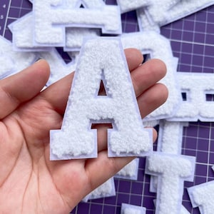 Könnte beinhalten: Weißer Filzbuchstabe A Patch. Der Patch ist flauschig und hat einen weißen Umriss. Der Buchstabe wird in einer Hand gehalten.
