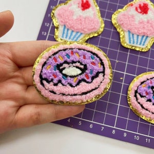 Puede incluir: Una dona rosa y morada con chispas de colores y un centro de glaseado blanco. La dona está rodeada por un borde de purpurina dorada.