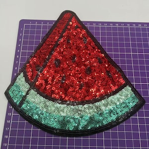 Può includere: Un cerotto a forma di anguria rosso e verde fatto di paillettes. L'anguria è tagliata a metà e ha un contorno nero.