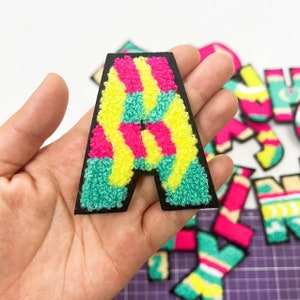May include: A colourful, fuzzy letter A patch with pink, yellow and green stripes. The patch is surrounded by other letters in a similar style.