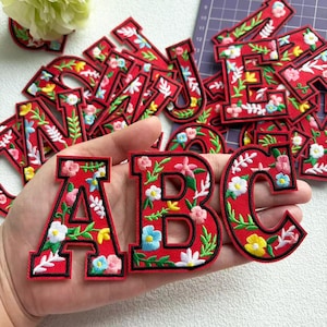Puede incluir: Letras rojas bordadas con diseños florales. Las letras A, B y C están enfocadas, con otras letras dispersas en el fondo. Las flores son de color rosa, azul, amarillo y blanco, con hojas verdes.