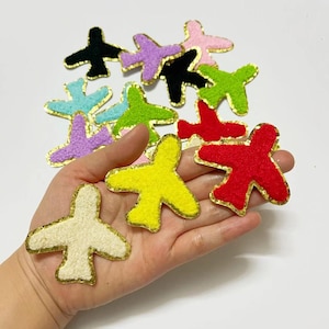May include: Assortment of colourful felt airplane patches with gold trim. The patches are in various colours, including red, yellow, green, blue, purple, and white.