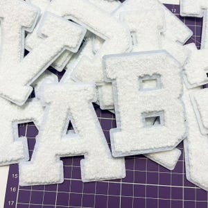 Op de afbeelding: Een stapel witte vilt letter patches, waaronder de letters A en B. De patches liggen op een paarse snijmat met raster.