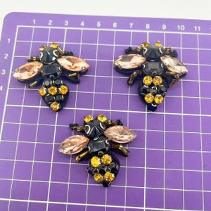 May include: Three black and gold bee-shaped embellishments with pink crystal wings. The bees are made of beads and rhinestones.
