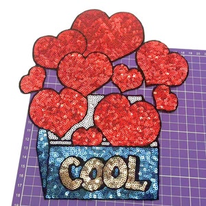 以下が含まれることがあります： 赤いスパンコールのハートのパッチで、「COOL」という文字が青と金の色のスパンコールで書かれています。パッチは、より小さな赤いスパンコールのハートで囲まれています。