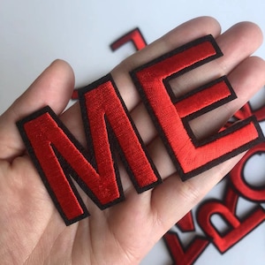 Può includere: Toppe di lettere in feltro rosso con contorni neri. Le lettere compongono la parola "ME".