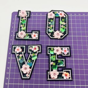 May include: Four felt letters spelling out the word "LOVE" in black with white outlines. Each letter is decorated with pink and green sequins and beads.