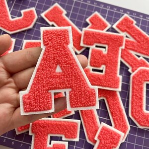 May include: Red chenille letter patches with white borders. The letter "A" is in focus, with other letters visible in the background. These patches are likely for customizing apparel or accessories. The patches have a textured, fuzzy appearance.