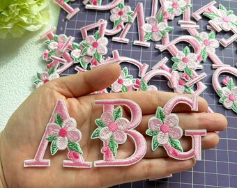 Könnte beinhalten: Gestickte Buchstaben A, B und G aus rosa Stoff, verziert mit rosa Blüten und grünen Blättern. Die Buchstaben werden vor einer lila Schneidematte gezeigt, mit anderen Buchstaben im Hintergrund. Die Buchstaben sind in einer Hand.
