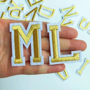 May include: Gold embroidered letters "M" and "L" on a white felt background. The letters are sewn with a white outline.