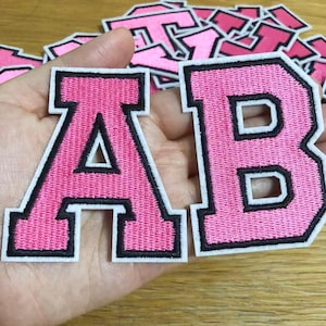 May include: Pink embroidered letter patches, including the letters "A" and "B", with a black border. The patches have a classic varsity style, perfect for customizing apparel or accessories. Other pink letter patches are visible in the background.