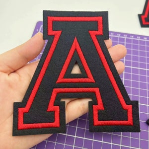 Puede incluir: Una letra A grande de fieltro negro con un contorno rojo. La letra mide aproximadamente 10 cm de alto y 7,5 cm de ancho.