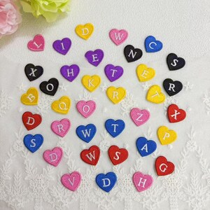 May include: A selection of heart-shaped fabric patches in various colours, each embroidered with a different letter of the alphabet. The patches are in shades of red, pink, yellow, purple, blue, and black. They are scattered on a white, textured surface.