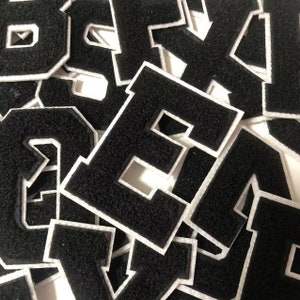 Puede incluir: Un montón de parches de letras de fieltro negro con contornos blancos. Las letras son de varios estilos, incluyendo letras de bloque y letras de escritura.