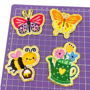 May include: Four embroidered iron-on patches with a gold glitter outline. The patches feature a pink and purple butterfly, an orange butterfly, a yellow bee with a pink flower, and a green watering can with flowers.