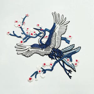 May include: Blue and white embroidered iron-on patch of a crane with outstretched wings perched on a branch of a flowering cherry blossom tree. The crane is facing left and the cherry blossom branch is facing right. The cherry blossoms are white with red centers.