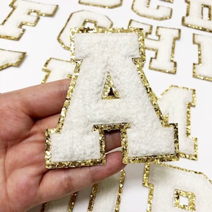 May include: White felt letter "A" with gold glitter trim. The letter is part of a set of alphabet patches. The patches have a fuzzy texture and are likely for decorative use on clothing or accessories.