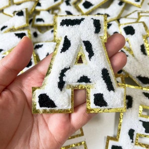 Könnte beinhalten: Ein weißer und schwarzer Kuhflecken-Buchstaben-A-Patch mit goldfarbenem Glitzerbesatz. Der Buchstabe ist aus flauschigem Stoff und perfekt, um Kleidung, Taschen oder anderen Accessoires eine persönliche Note zu verleihen.