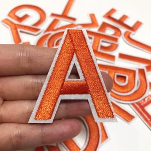 Puede incluir: Letra A de fieltro naranja con contorno blanco. La letra está bordada con hilo naranja y tiene un respaldo blanco.