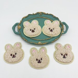 May include: Five beige fluffy bunny face patches with pink ears and brown eyes. The patches are on a teal tray with gold trim.