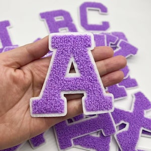 May include: Purple felt letter patches in the alphabet, with a white outline. The letter A is being held in a hand.