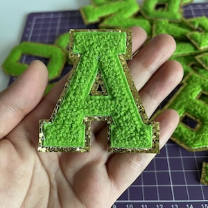 以下が含まれることがあります： 金色のグリッターの縁取りが施された緑色のフェルトの文字A。文字の高さは約5cm、幅は約4cmです。