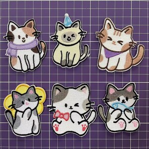 May include: Six embroidered iron-on patches featuring cute cartoon cats in various poses. The cats are white with brown, gray, or orange markings. Some cats are wearing accessories like a scarf, a party hat, or a bow tie.