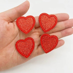 May include: Four red glitter heart-shaped patches. The hearts are outlined in red thread.