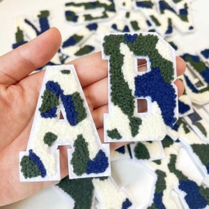 May include: A set of felt letters in a camouflage pattern. The letters are white with green and blue camouflage print. The letters are A and B.