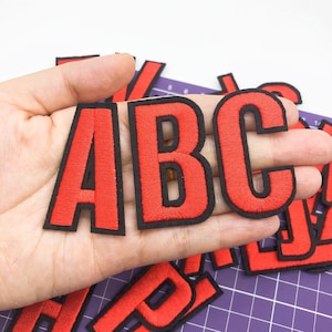 Pode incluir: Letras de feltro vermelho com contornos pretos, soletrando "ABC". As letras são patches termocolantes, perfeitas para adicionar um toque personalizado a roupas, bolsas ou outros itens de tecido.