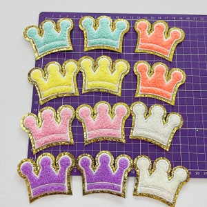 Pode incluir: Dez patches de coroa de feltro com acabamento dourado de glitter. As coroas são de várias cores, incluindo azul claro, amarelo, laranja, rosa, roxo e branco.