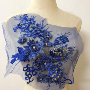 以下が含まれることがあります： 詳細な刺繍が施された青い花のアップリケ。デザインは、青い花、シルバーのアクセント、きらびやかな装飾が施された、透明な青いメッシュの裏地が特徴です。衣類を飾るのに最適です。