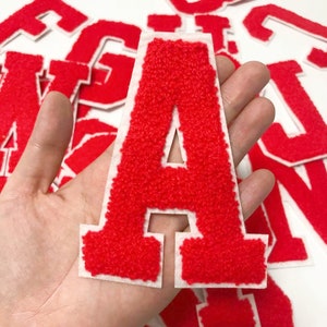 Puede incluir: Letra A de fieltro rojo con contorno blanco. La letra se sostiene en una mano sobre un fondo blanco.