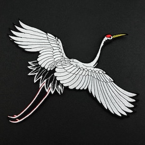 Op de afbeelding: Witte geborduurde kraanvogel met rode en gele accenten op een zwarte achtergrond. De kraanvogel is in vlucht met zijn vleugels wijd uitgespreid.
