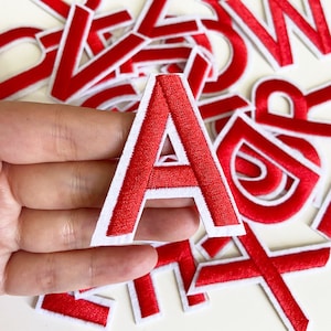 以下が含まれることがあります： 白い縁取りのある赤いフェルトの文字パッチ。Aの文字が手で持たれています。