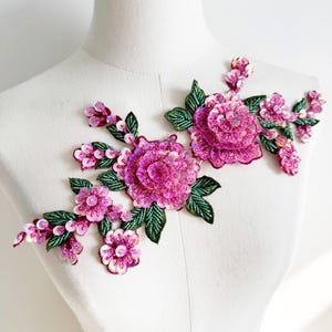 Puede incluir: Una aplicación floral que presenta dos grandes flores rosas con lentejuelas e iridiscencias, rodeadas de pequeños capullos rosas con lentejuelas y hojas bordadas en verde oscuro. El diseño está dispuesto en un patrón horizontal ramificado.