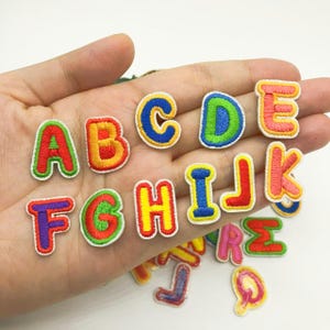 May include: A set of colourful embroidered iron-on alphabet patches. The patches are in various colours, including red, yellow, blue, green, and orange. Each patch features a single letter of the alphabet in a bold, outlined font.