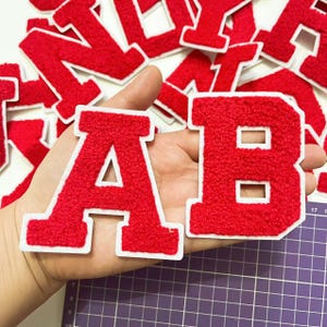 May include: Red felt letter patches with white outlines. The letters are A and B. The patches are held in a hand.