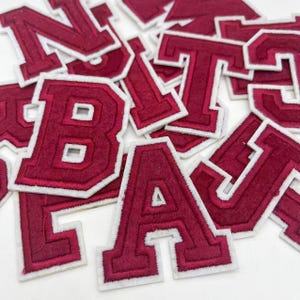 Puede incluir: Un conjunto de letras de fieltro burdeos con contornos blancos. Las letras están en mayúsculas e incluyen las letras A, B, J, L, N, T y U.