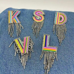 Op de afbeelding: Vijf kleurrijke regenboog-letterpatches met strass franjes. De letters zijn K, S, D, V en L. De letters zijn genaaid op een denim achtergrond.