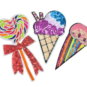 Puede incluir: Tres parches termoadhesivos coloridos que representan un cono de helado rosa y azul, un cono de helado rosa con rayas arcoíris y una piruleta en forma de corazón rojo y blanco con un lazo rojo.