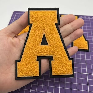 以下が含まれることがあります： 黒い輪郭のある大きな黄色のフェルトの文字A。文字は厚みがあり、ふわふわした質感です。