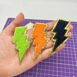 May include: Three lightning bolt shaped patches with gold glitter trim. The patches are made of fuzzy fabric and are in green, orange, and black.