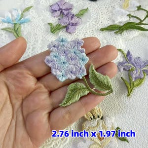 以下が含まれることがあります： 青、紫、白など、さまざまな色の刺繍された花のパッチ。 パッチは詳細なステッチと緑の葉が特徴です。 画像のテキストは「2.76インチx 1.97インチ」（7.01 cm x 5.00 cm）です。