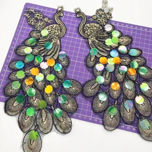 Könnte beinhalten: Zwei goldene gestickte Pfauenmotive mit schillernden Pailletten in Grün-, Blau- und Orangetönen. Die Motive befinden sich auf einem violetten, karierten Hintergrund.